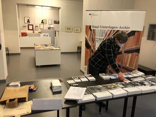 Stasi-Unterlagen-Archiv Cottbus (im Aufbau) Eine Mitarbeiterin des Stasi-Unterlagen-Archivs steht hinter einem Tisch mit Informationsbroschüren. Im Hintergrund ist ein Rollup des Stasi-Unterlagen-Archivs zu sehen.