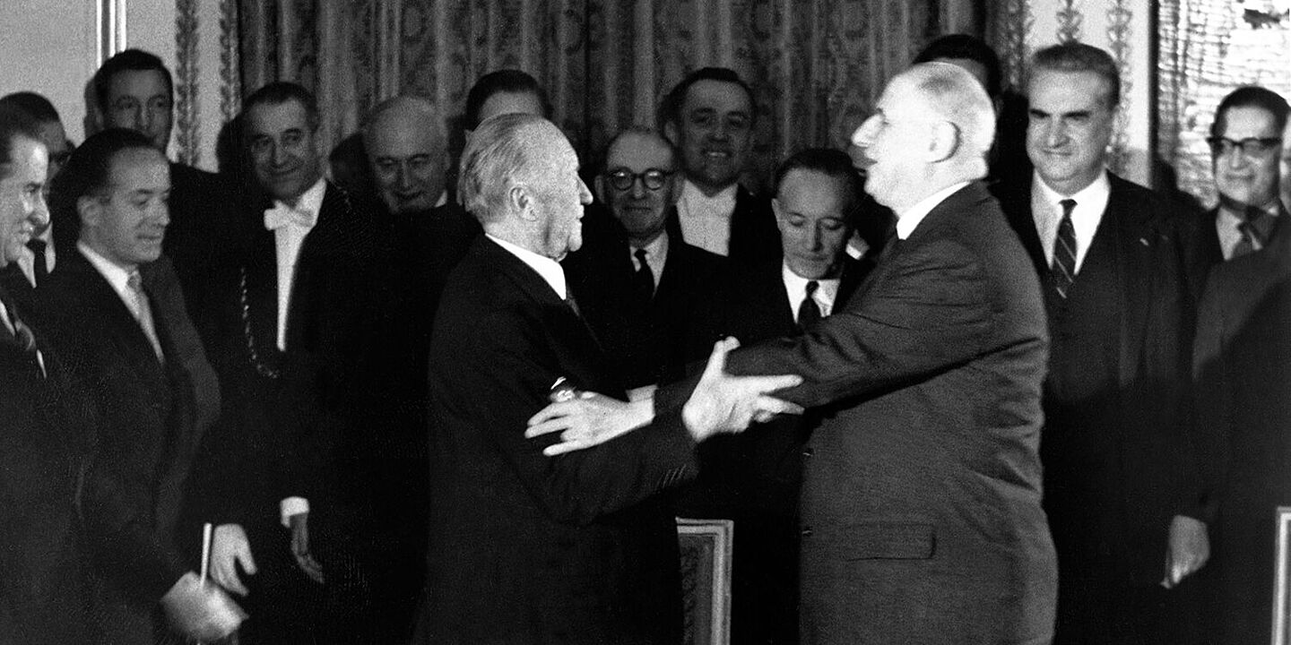 Konrad Adenauer und Charles de Gaulle im Élysée-Palast 1963 Konrad Adenauer und Charles de Gaulle strecken ihre Arme zueinander aus, um sich zu umarmen. Im Hintergrund stehen mehrere Menschen, die lächeln., Quelle:
BArch, B 145 Bild-00361399 / Schwahn, Ernst