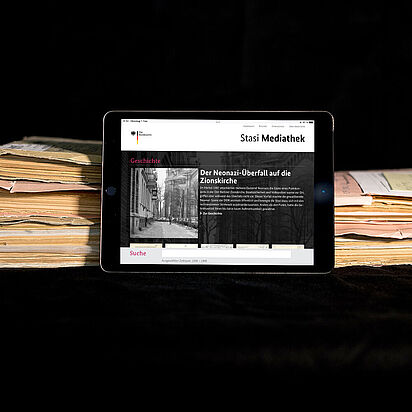 Mediathek Das Bild zeigt die auf einem Tablet geöffnete Startseite der Stasi-Mediathek. Das Tablet lehnt an einigen Aken.