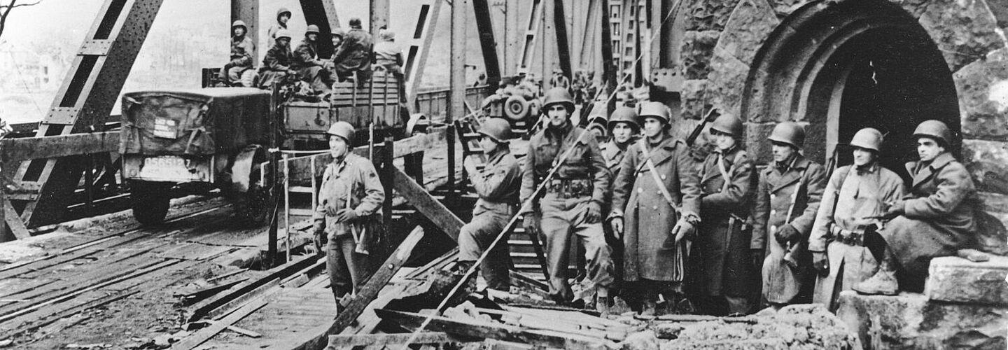 Amerikanische Soldaten auf der Brücke von Remagen zwischen dem 8. und 10. März 1945 Mehrere amerikanische Soldaten stehen auf der beschädigten Brücke von Remagen. Links sind ein Jeep und ein LKW mit Anhänger zu sehen, auf dem ebenfalls Soldaten sitzen.