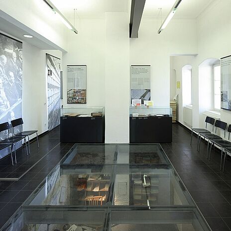 Dauerausstellung in Ludwigsburg Blick in die Dauerausstellung des Bundesarchivs im Schorndorfer Torhaus in Ludwigsburg. Zu sehen sind verschiedene Glasvitrinen, mehrere Informationstafeln und Stühle.