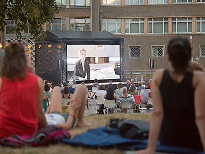 Gemütlich gemacht vor historischer Kulisse: Campus-Kino. Gemütlich gemacht vor historischer Kulisse: Menschen schauen auf eine Leindwand im Innenhof der ehemaligen Stasi-Zentrale.