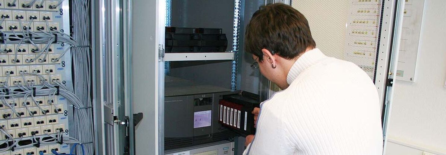 Ausbildung Fachinformatiker/in Eine Person steht vor einem IT-System in einem Schrank und holt etwas aus dem Schrank raus.