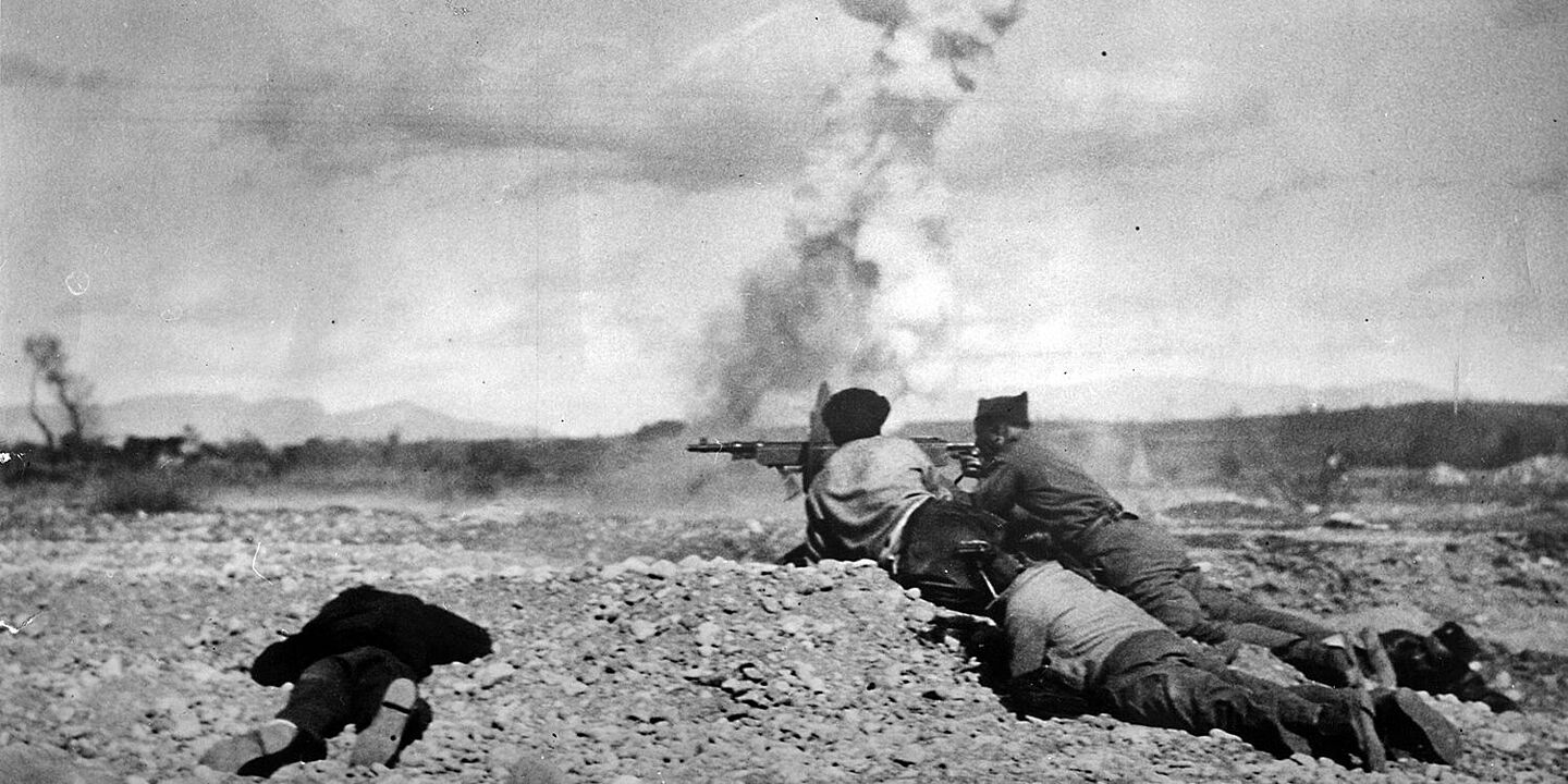 Republikanische Soldaten in Spanien 1937 Schwarz-Weiß-Aufnahme von vier Männern, die mit Gewehren im Anschlag auf und hinter einem Schotterhügel liegen. Im Hintergrund ziehen hohe Rauchschwaden in den Himmel., Quelle:
BArch, Bild 183-K1013-501 / o. Ang.