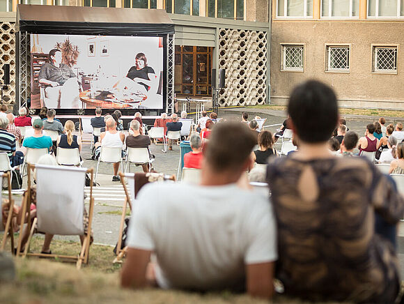 Campus-Kino in der ehemaligen Stasi-Zentrale Zuschauer sitzen auf Stühlen und auf der Wiese und schauen in Richtung einer großen Leinwand.