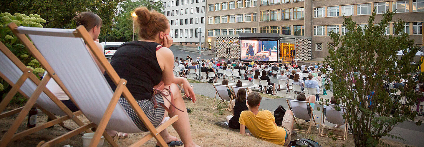 Impression vom Campus-Kino 2020 Menschen sitzen im Innenhof der ehemaligen Stasi-Zentrale auf Stühlen und Liegestühlen und schauen auf eine Kinoleinwand im Hintergrund.