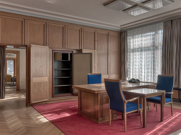 Mielke-Büro Das Bild zeigt ein hochwertig eingerichtetes Büro mit Holzverkleidung. In der Mitte des Raumes steht ein großer Schreibtsich mit drei Stühlen. Die Fenster sind mit halbdurchlässigen Vorhängen abgedeckt