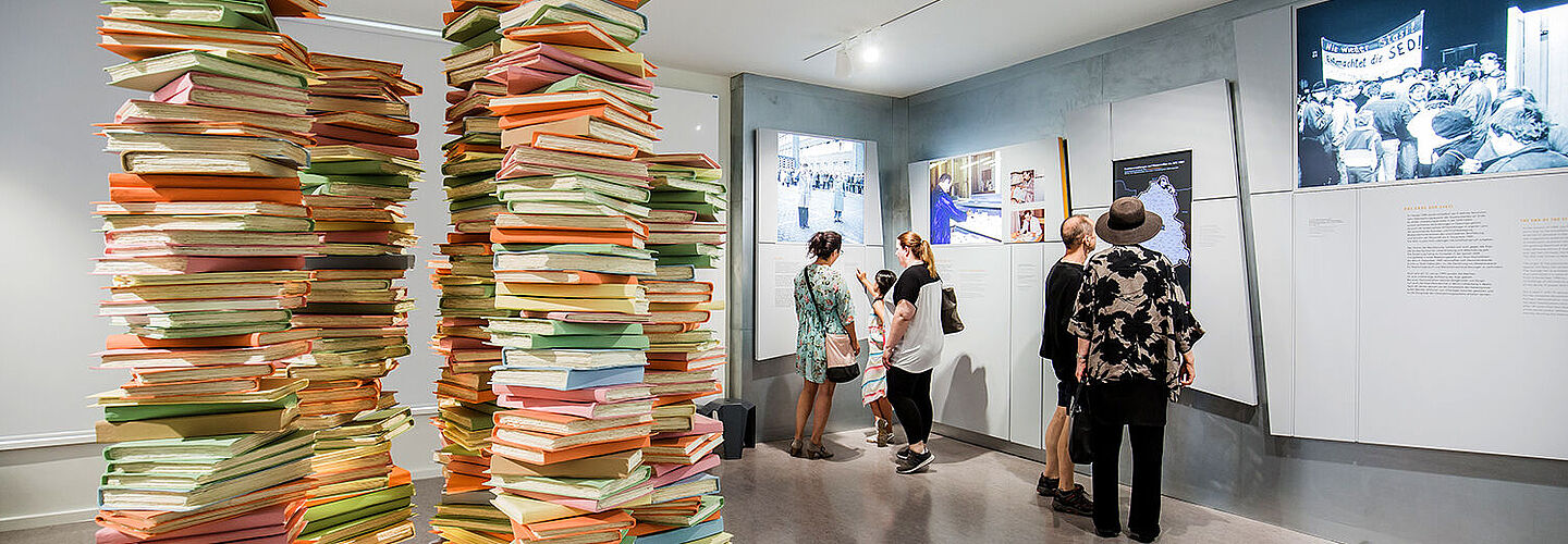 Blick in die Dauerausstellung „Einblick ins Geheime“ in Berlin-Lichtenberg Das Bild zeigt im Vordergrund eine Akteninstallation, Im Hintergrund einige Besucher an verschiedenen Medienstationen.